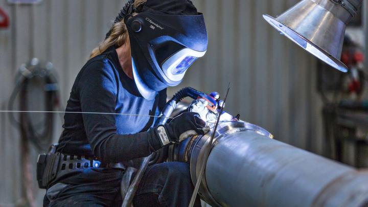 Woman welding a metal pipe with a protective helmet and gloves, illuminated by bright light in a workshop setting.