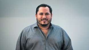 Yosafat Esquitin, Business Development Manager, standing against a plain light background wearing a patterned button-up shirt.