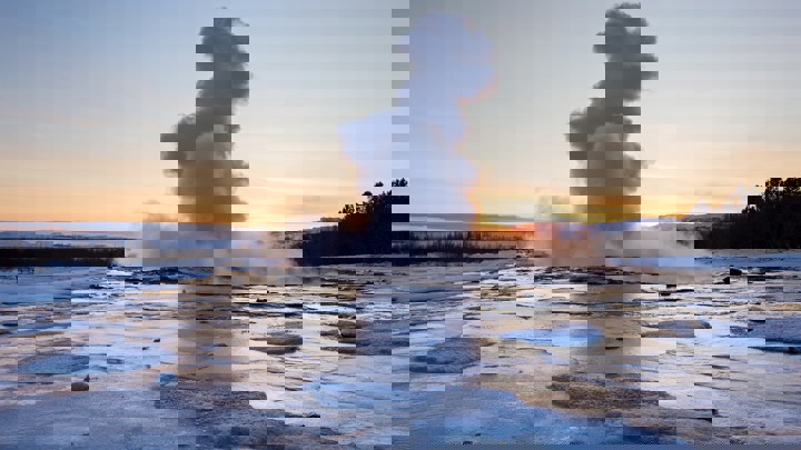 A steam plume rises from a geyser against a sunset, surrounded by icy landscapes and snow-covered trees visible in the distance.